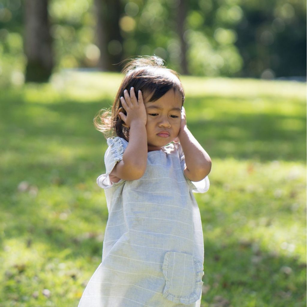 Small kid wearing white dress in park covering her ears in ADHD frustration