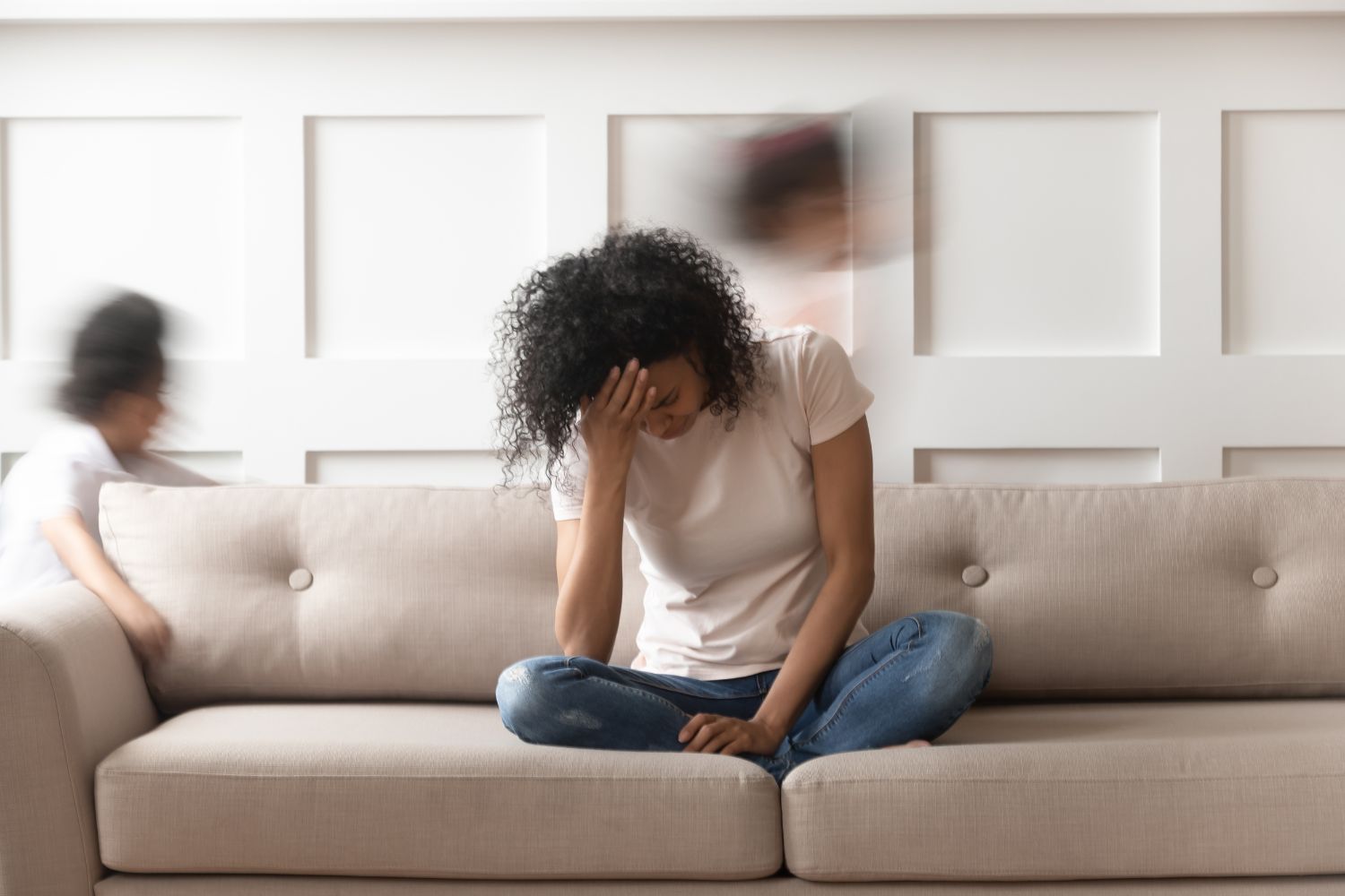 MOM ON GREY SOFA WITH HEAD IN HANDS IN PARENTING GUILT-KID RANS AROUND